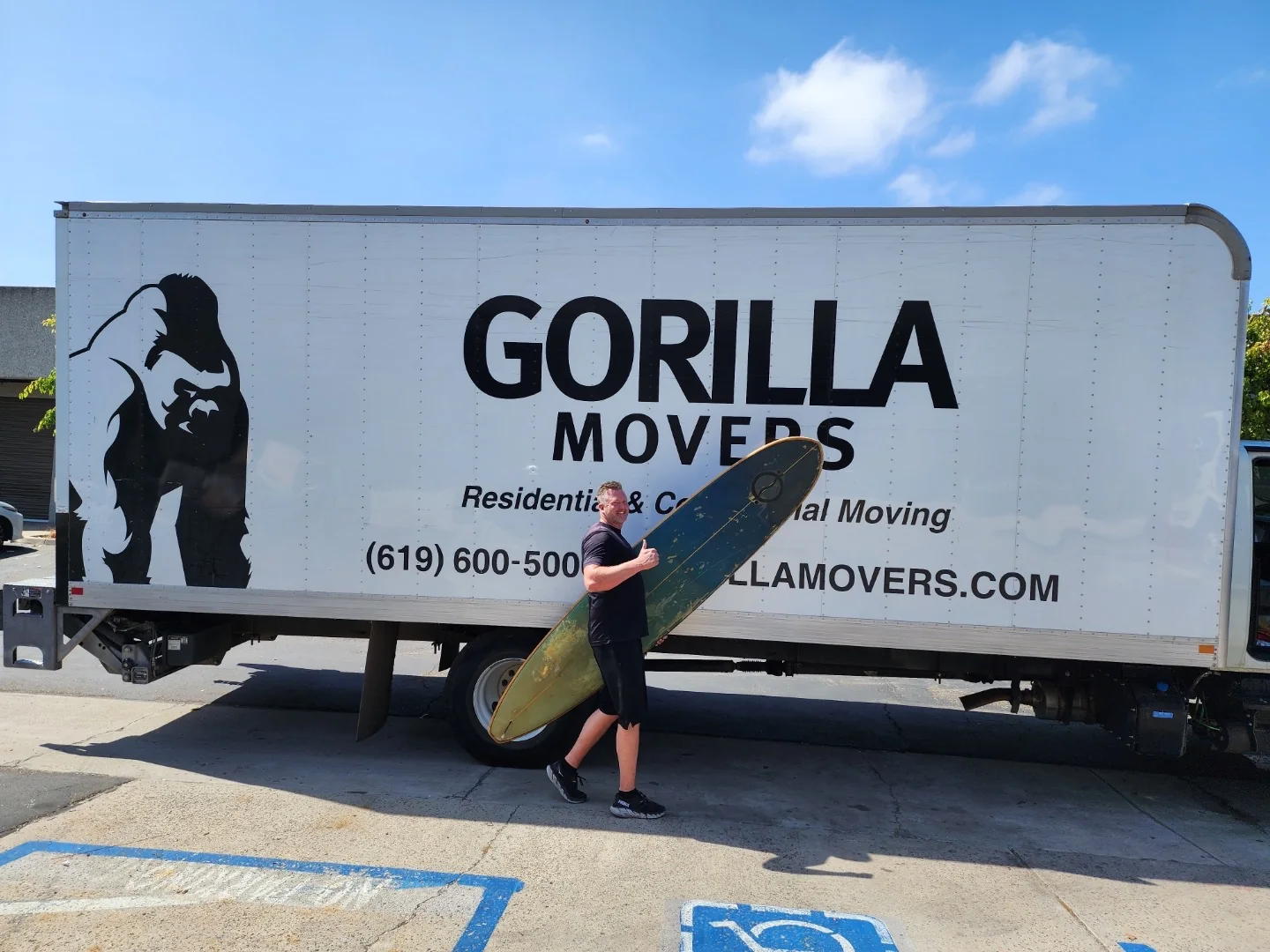 Gorilla Movers owner carrying a longboard surfboard past a branded Gorilla Movers box truck in San Diego